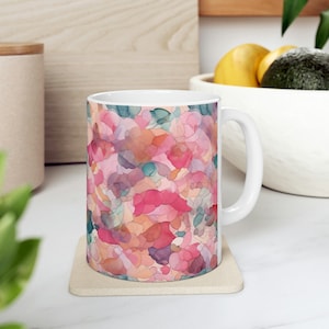 May include: White ceramic mug with a pink, orange, and blue abstract floral pattern. The mug is sitting on a beige coaster.
