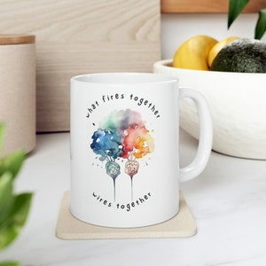 May include: White ceramic mug with a watercolor design of two trees with rainbow colored leaves. The text "what fires together wires together" is printed on the mug.