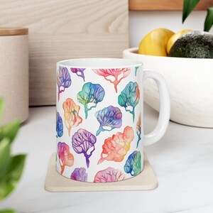 Op de afbeelding: Witte keramische mok met een kleurrijk aquarelpatroon van bomen. De bomen zijn in tinten roze, blauw, groen, paars en oranje.