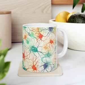 Op de afbeelding: Witte keramische mok met een kleurrijk aquarelontwerp van neuronen. Het ontwerp bevat een verscheidenheid aan kleuren, waaronder blauw, groen, oranje en rood.
