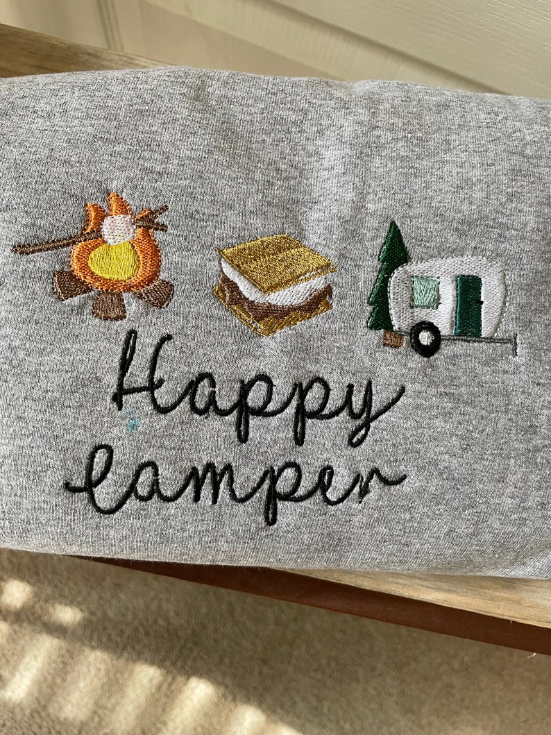 May include: Grey sweatshirt with embroidered images of a campfire, a s'more, and a camper van. The text "Happy Camper" is embroidered below the images.