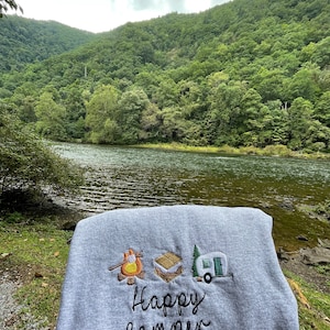 May include: Grey sweatshirt with embroidered camping icons, including a campfire, a marshmallow, and a camper van. The text "Happy Camper" is embroidered below the icons.