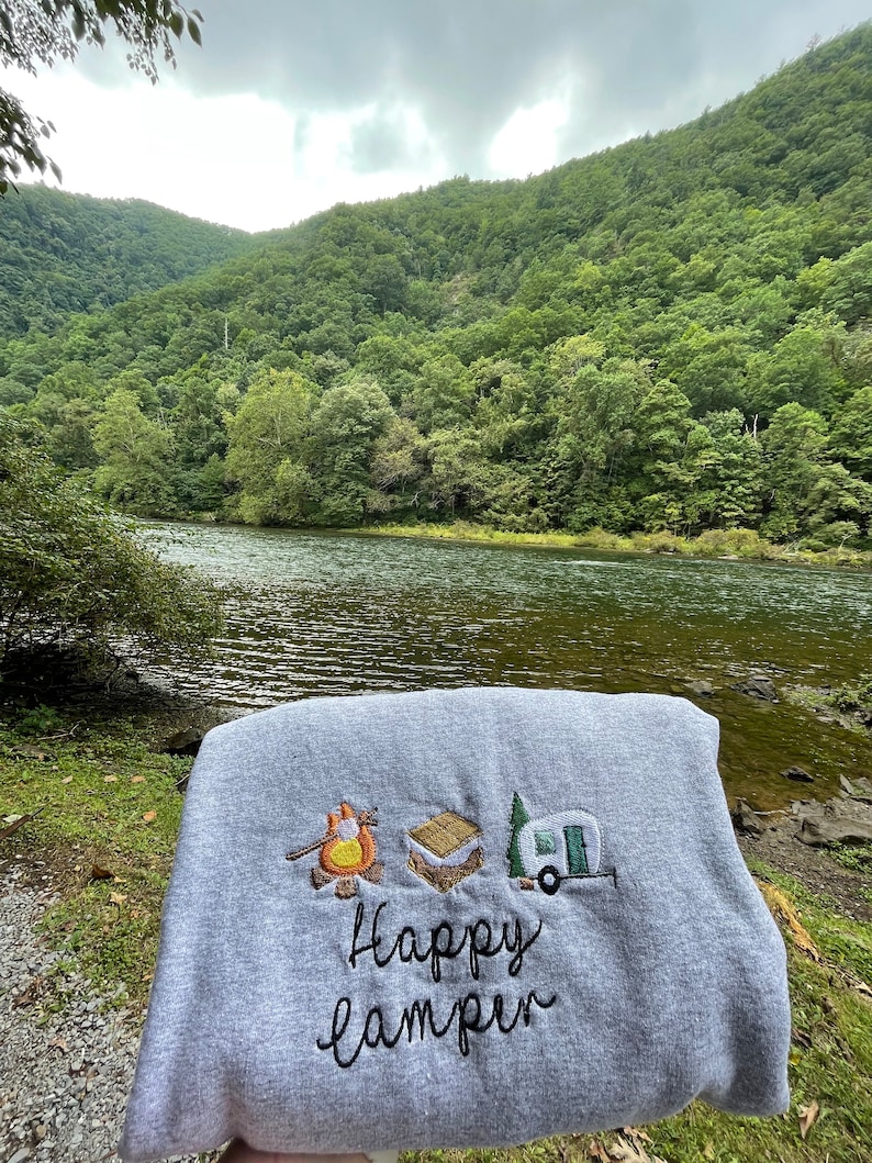 May include: Grey sweatshirt with embroidered camping icons, including a campfire, a marshmallow, and a camper van. The text "Happy Camper" is embroidered below the icons.
