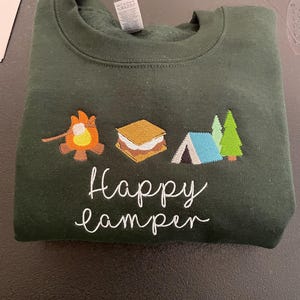 May include: A dark green sweatshirt with embroidered images of a campfire, a marshmallow, a tent, and trees. The text "Happy Camper" is embroidered in white below the images.
