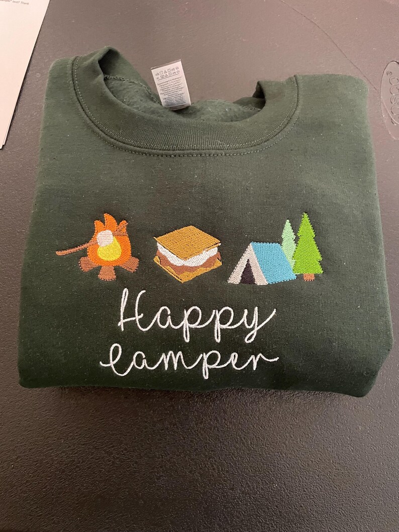 May include: A dark green sweatshirt with embroidered images of a campfire, a marshmallow, a tent, and trees. The text "Happy Camper" is embroidered in white below the images.