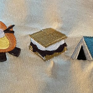 May include: Embroidered design of a campfire with a marshmallow on a stick, a s'more, a tent, and two pine trees. The design is stitched on a white fabric.