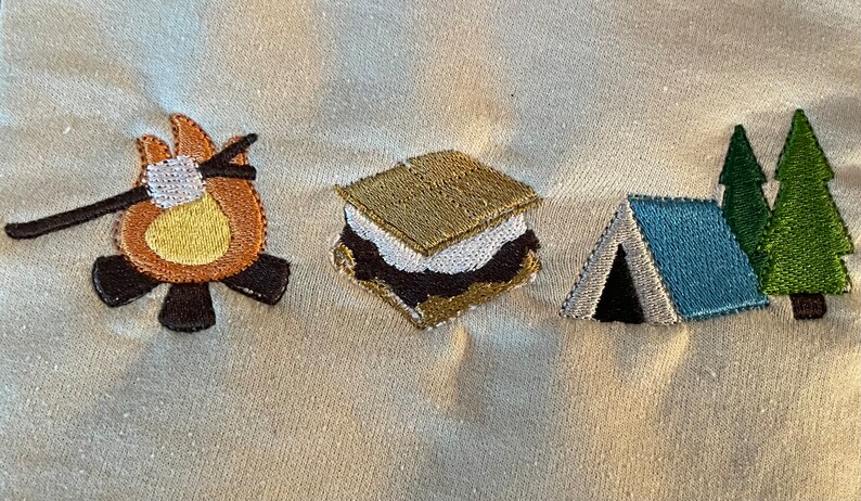 May include: Embroidered design of a campfire with a marshmallow on a stick, a s'more, a tent, and two pine trees. The design is stitched on a white fabric.