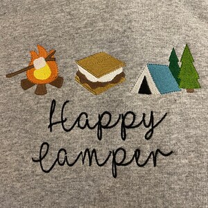 May include: Grey sweatshirt with embroidered camping icons, including a campfire, a s'more, a tent, and two pine trees. The text "happy camper" is embroidered below the icons.