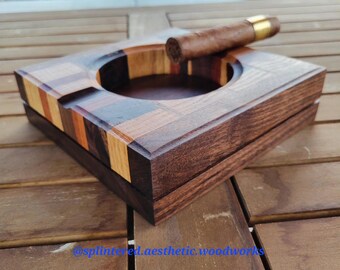 May include: A square wooden ashtray with a cigar resting on the edge. The ashtray is made of various wood tones, including dark brown, light brown, and orange. The ashtray has a circular indentation for holding the cigar and is sitting on a wooden surface.