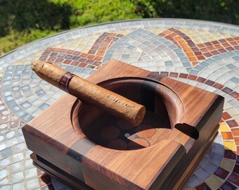 May include: A square wooden ashtray with a cigar resting in it. The ashtray is made of dark wood with a circular indentation for the cigar and a small notch. The cigar has a brown wrapper and a red and gold band.