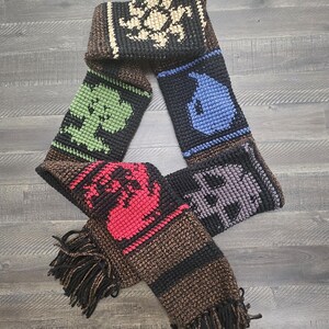 May include: A handmade crochet scarf featuring various colorful geometric designs. The scarf has a brown, black, and cream color scheme with fringe at the ends. The scarf is made of yarn and is displayed on a wooden surface.