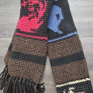 May include: A handmade crochet scarf with a brown and black base, featuring red and blue geometric designs. The scarf has black fringe at the ends. The scarf is made of soft yarn and is suitable for cold weather.