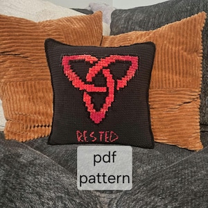 May include: A black cushion featuring a red Celtic knot design and the word "RESTED" embroidered in red. The cushion is set against a backdrop of brown corduroy cushions and a grey blanket. The text "pdf pattern" is in a white rectangle.