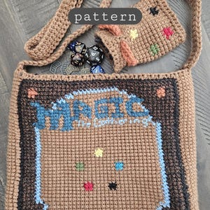 Könnte beinhalten: Gehäkelte braune Umhängetasche mit dem Text "MAGIC the Gathering" und "DECKMASTER" in Blau und Grün. Enthält eine passende kleine Tasche und Würfel. Die Tasche hat einen braunen Riemen und ist aus strukturiertem Garn gefertigt.