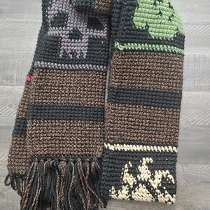 May include: A handmade crochet scarf in shades of black, brown, and gray, featuring skull and leaf motifs. The scarf has a fringed edge and is displayed on a wooden surface. The design includes stripes and textured patterns.