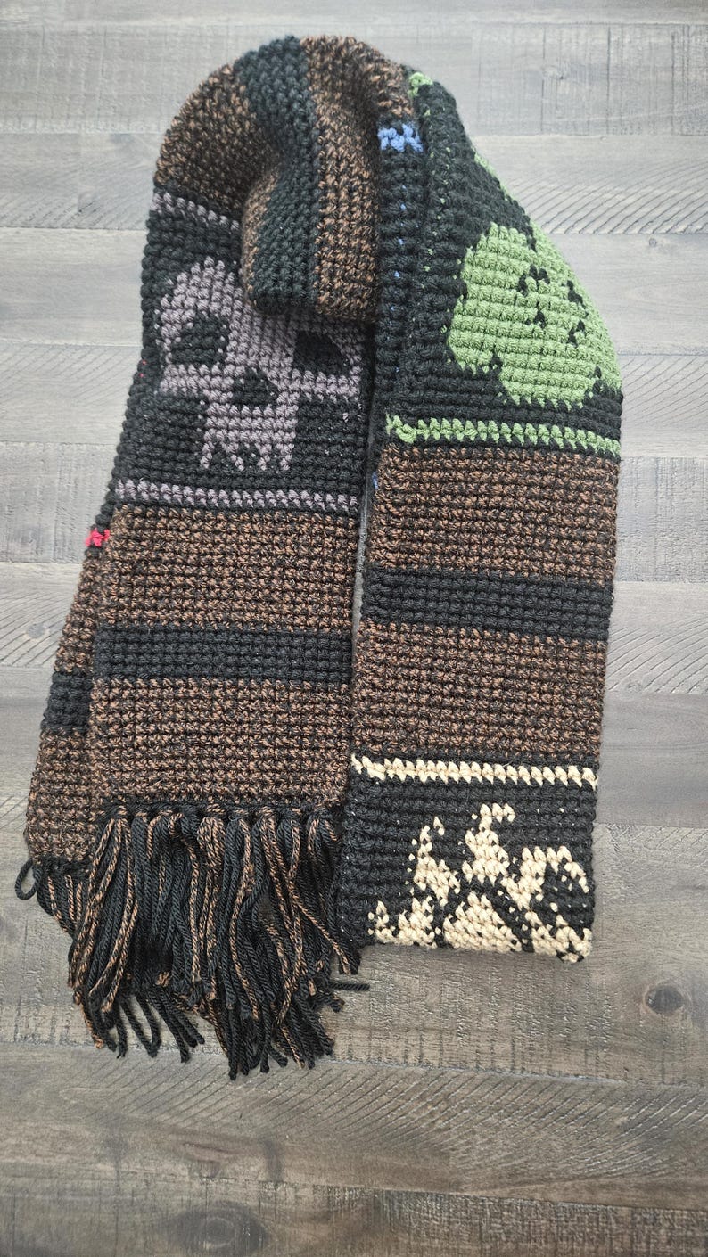 May include: A handmade crochet scarf in shades of black, brown, and gray, featuring skull and leaf motifs. The scarf has a fringed edge and is displayed on a wooden surface. The design includes stripes and textured patterns.