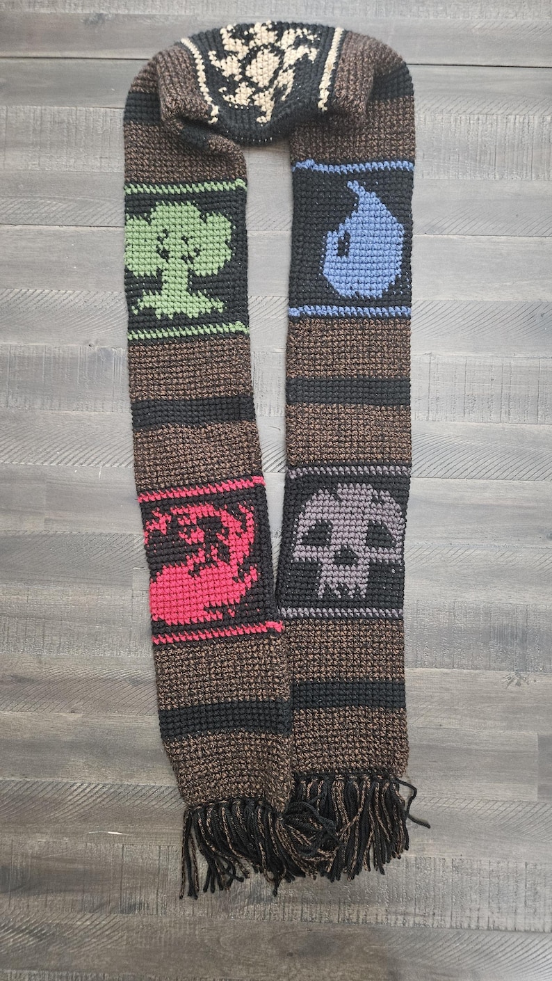 May include: A brown and black crocheted scarf with various symbols in black, green, red, blue, and gray. The scarf has a fringe at the bottom. The symbols include a tree, a skull, and other abstract designs.