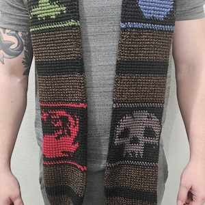 May include: Crocheted scarf with a brown base and black stripes, featuring green, blue, red, and gray symbols. The scarf has a fringed bottom. The symbols include a tree, a water drop, a dragon, and a skull.