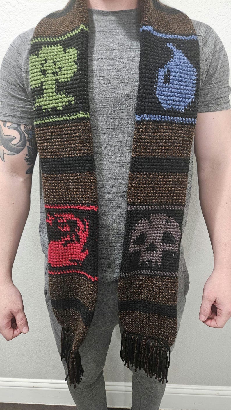 May include: Crocheted scarf with a brown base and black stripes, featuring green, blue, red, and gray symbols. The scarf has a fringed bottom. The symbols include a tree, a water drop, a dragon, and a skull.