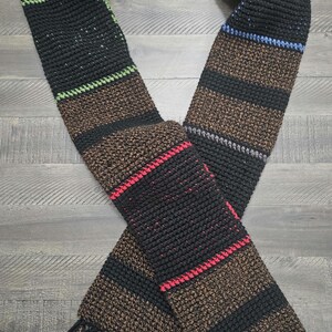 May include: A handmade scarf with a striped pattern in black, brown, and red. The scarf has fringe at the ends and is made of a textured yarn. Green, blue, and gray stripes accent the design. A cozy winter accessory.