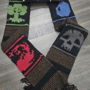 May include: A handmade crochet scarf featuring various graphic designs. The scarf has a brown base with black and colored accents. Designs include a snowflake, tree, blue alien, skull, and a red creature. The scarf has a fringed edge.