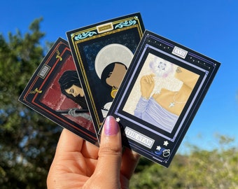 以下が含まれることがあります： 黒い背景と金のアクセントが付いたタロットカード3枚。カードは「星」「月」「死神」と題されています。各カードには、それぞれ異なる人物のイラストが描かれています。