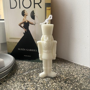 May include: White ceramic nutcracker candle with a white string wick. The candle is shaped like a traditional nutcracker with a hat, mustache, and a red and white striped uniform.