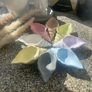 Puede incluir: Un conjunto de nueve conchas de cerámica de color pastel, cada una con una forma y un diseño únicos. Las conchas están dispuestas en círculo sobre una superficie gris.