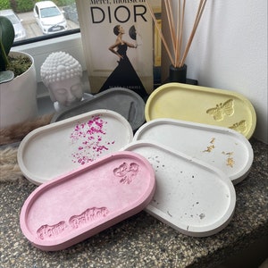 Puede incluir: Cinco bandejas ovaladas de hormigón en diferentes colores. Las bandejas tienen varios diseños, incluyendo una mariposa, una flor y las palabras "Feliz cumpleaños".
