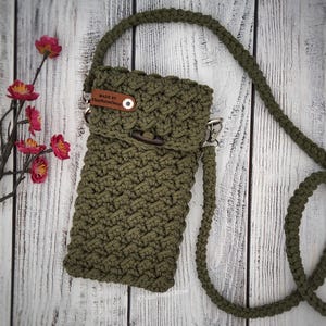May include: An olive green crossbody bag crafted with a textured macrame knot pattern. The bag features a long matching shoulder strap, a fold-over flap with a dark wooden toggle closure, and a small brown leather tag labeled MADE BY ViasHomeDecor.