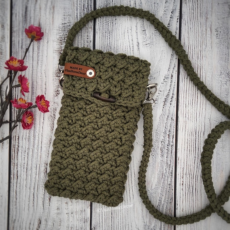 May include: An olive green crossbody bag crafted with a textured macrame knot pattern. The bag features a long matching shoulder strap, a fold-over flap with a dark wooden toggle closure, and a small brown leather tag labeled MADE BY ViasHomeDecor.