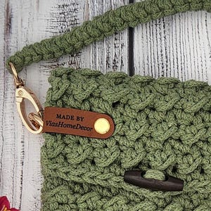 May include: A sage green crocheted bag featuring a thick braided strap attached with a gold-toned metal lobster clasp. A brown leather tag embossed with the text MADE BY ViasHomeDecor is secured with a gold rivet, and a dark wooden toggle closure.