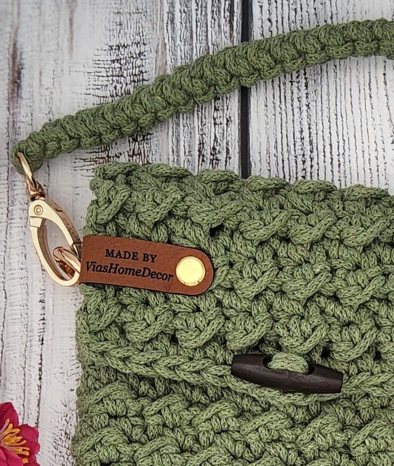May include: A sage green crocheted bag featuring a thick braided strap attached with a gold-toned metal lobster clasp. A brown leather tag embossed with the text MADE BY ViasHomeDecor is secured with a gold rivet, and a dark wooden toggle closure.