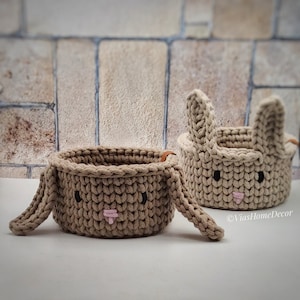 Könnte beinhalten: Zwei beigefarbene gehäkelte Körbe in Hasenform mit braunen Ledergriffen. Die Körbe haben schwarze Augen und eine rosa Nase.