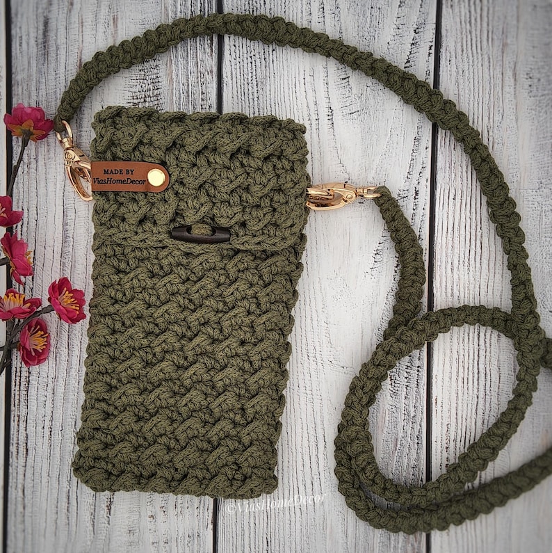 May include: An olive green crocheted crossbody bag featuring a textured stitch pattern, a long matching strap, and gold-toned metal hardware. A small brown leather tag is attached to the side with the text "MADE BY ViasHomeDecor" printed on it.