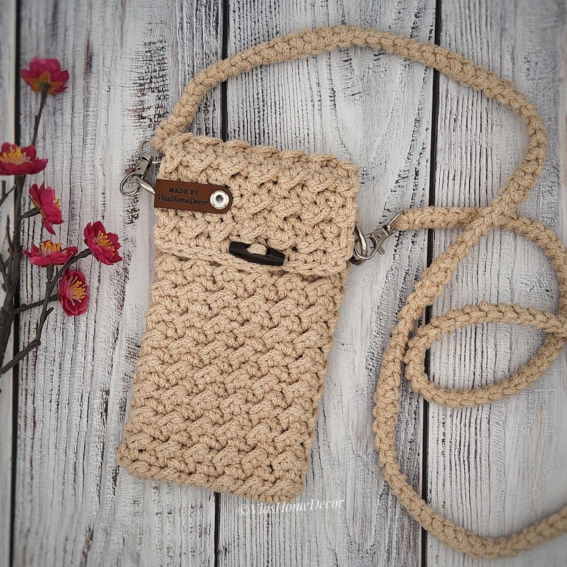 May include: A beige crochet crossbody bag with a textured pattern, a long matching strap, and a flap closure secured by a dark brown button. A small brown leather tag on the flap features the text Made By ViasHomeDecor in white lettering.