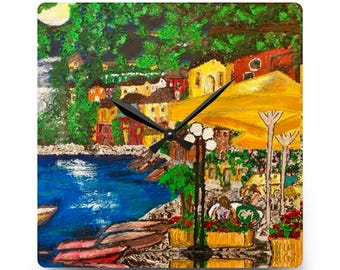 May include: Square wall clock with a vibrant painting of a coastal town. The artwork depicts a blue sea, yellow parasols, and buildings in yellow, red, and brown. Black clock hands show the time. A decorative item for the home.