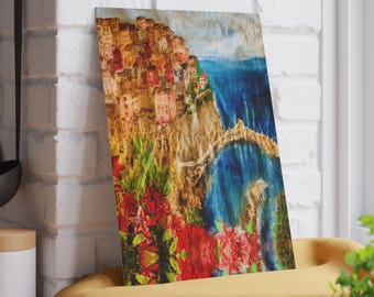 May include: A rectangular art print showing a coastal village with vibrant buildings on a cliff. The image includes a bridge over the blue sea, with red flowers in the foreground. The print is propped against a white brick wall.