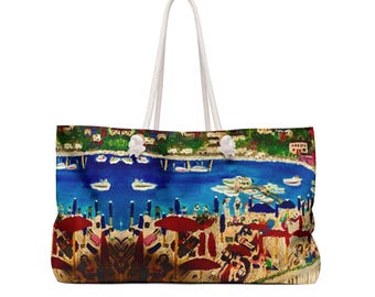 May include: Large tote bag with a colourful beach scene print. The design features a blue ocean, boats, red umbrellas, and a coastal town. The bag has white rope handles.
