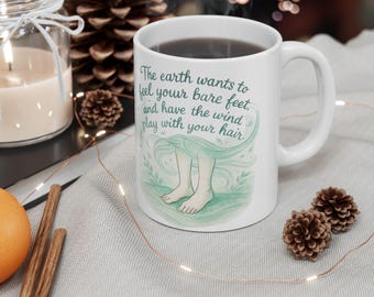 May include: White ceramic mug with a curved handle, filled with a dark liquid. The mug features the text "The earth wants to feel your bare feet, and have the wind play with your hair" and an illustration of feet. Decorative items surround the mug.