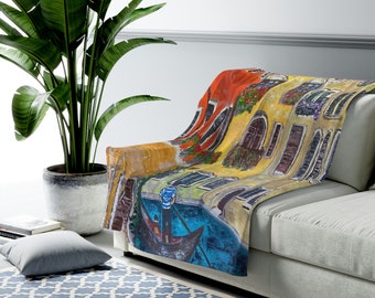 May include: A colourful throw blanket featuring a painting of a gondola in a canal with buildings on either side. The buildings are painted in shades of yellow, orange, and red, with green plants and flowers growing on the balconies. The gondola is blue and white, and the water is a deep blue.