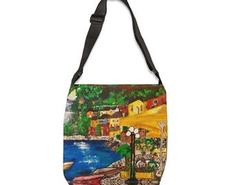 May include: A shoulder bag featuring a painting of a coastal scene. The artwork shows buildings in yellow, red, and orange, with a blue sea and green trees. The bag has a black adjustable strap.