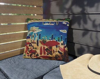 May include: A colourful throw pillow with a beach scene featuring people, boats, and umbrellas. The pillow is on a blue denim couch with a straw hat.