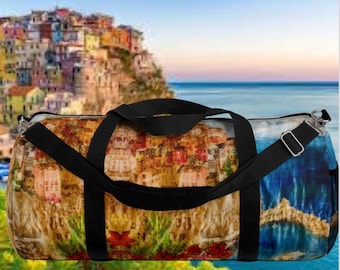 May include: A duffel bag with a vibrant print of a coastal town, complete with a black shoulder strap and handles. The bag has a zip closure and a side pocket. The background shows a coastal view.