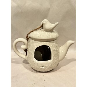 May include: A cream-colored ceramic teapot-shaped birdhouse with a glossy finish. The exterior features a raised floral vine pattern, a central arched opening, a curved handle, a spout, and a lid topped with a small sculpted bird figure.