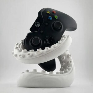 Op de afbeelding: Een zwarte Xbox-controller wordt vastgehouden door een witte octopusvormige standaard. De controller heeft gekleurde knoppen en een D-pad. De standaard heeft een gedetailleerd ontwerp met zuignappen. Een uniek gaming accessoire.