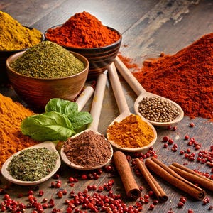 May include: A variety of spices in bowls and spoons, including turmeric, paprika, chili powder, coriander, and cinnamon sticks. The spices are arranged on a wooden surface with red peppercorns scattered around.