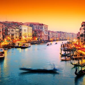 May include: A view of a canal in Venice, Italy, at sunset. The canal is lined with buildings and there are gondolas and other boats in the water. The sky is a vibrant orange and pink.