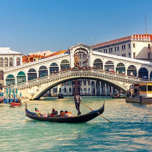 May include: A gondola with a gondolier in a white shirt and black pants is on a canal in Venice, Italy. The Rialto Bridge is in the background. The bridge is white with arches and is lined with shops and restaurants.