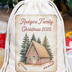 May include: A white drawstring bag featuring a rectangular graphic with the text Rodgers Family Christmas 2026. The graphic depicts a small cabin with red and white vertical stripes surrounded by green pine trees and two candy canes at the base.